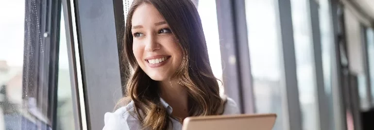 Smiling woman holding a tablet, gazing out of a large window.